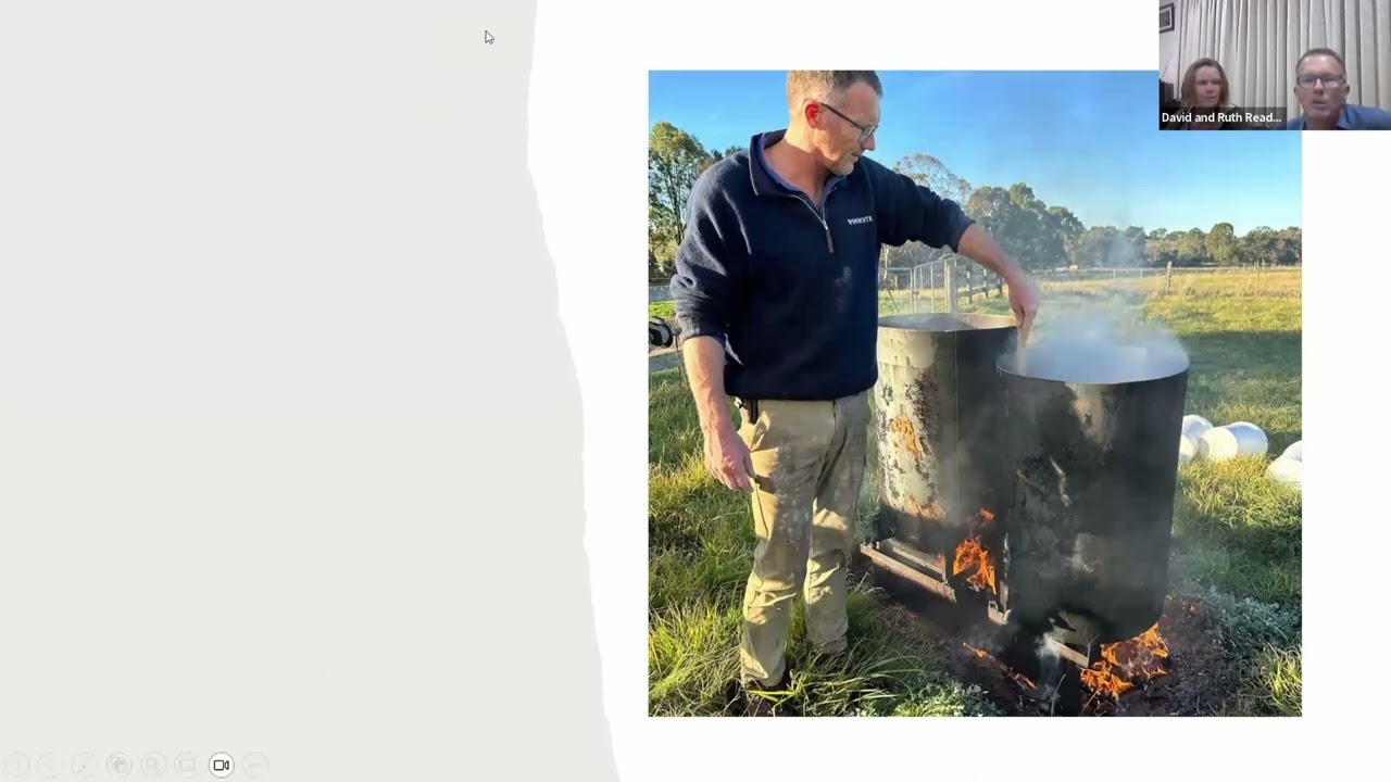 A man stands outdoors, attending to a large, steaming barrel with fire underneath it. He wears glasses and a navy blue sweater. In the top right corner, a small video thumbnail shows two people on a video call. Green grass and trees are visible in the background.