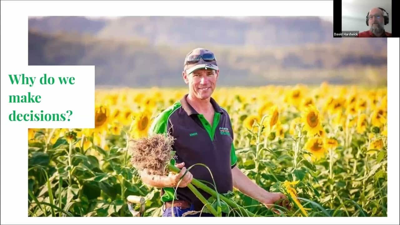 A farmer, dressed in a green and black shirt and cap, stands in a field of sunflowers holding a plant with exposed roots. Text on the left reads, "Why do we make decisions?" There is a small inset of a person in the top right corner.