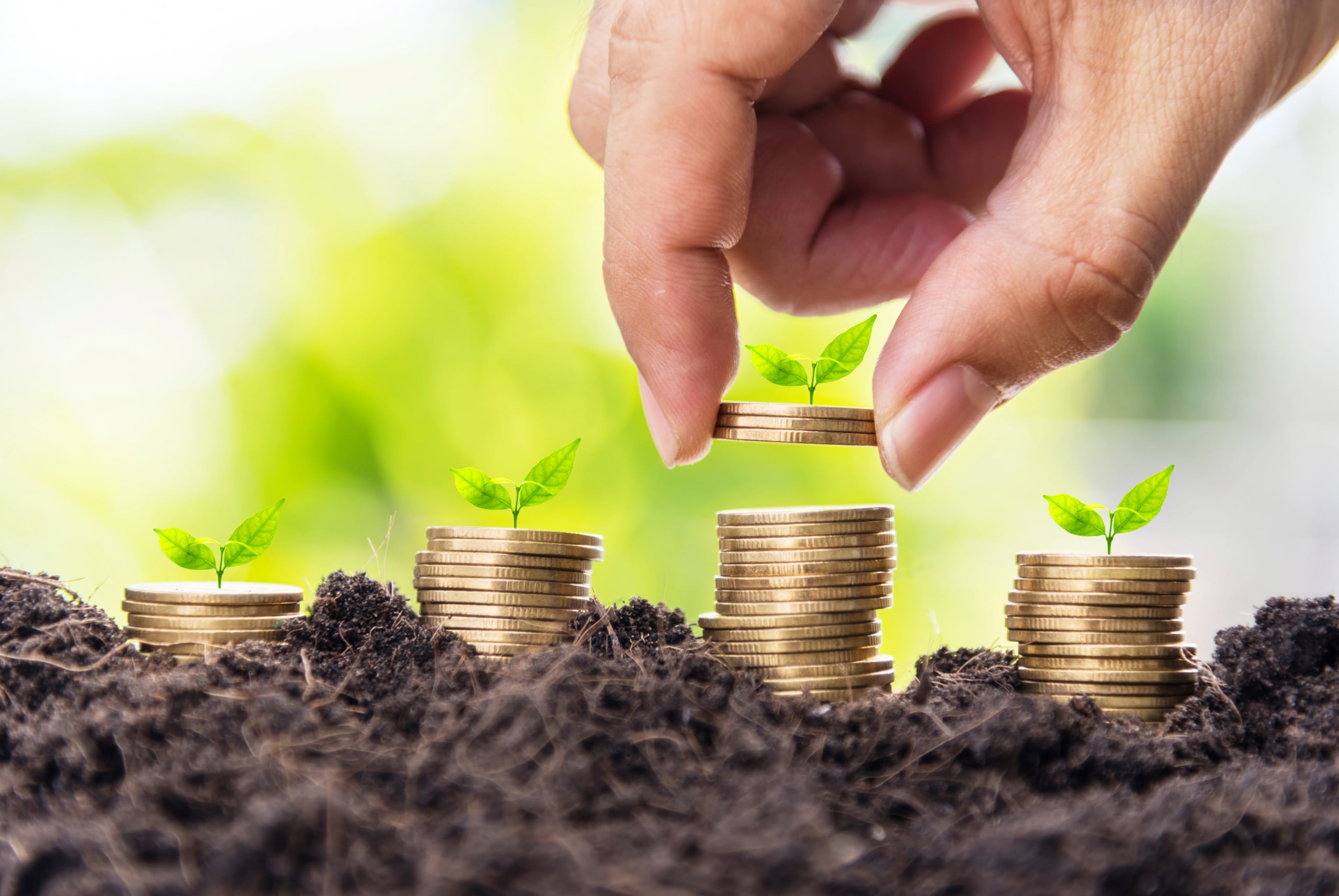 A hand is placing a gold coin on top of several stacks of coins, each topped with small green plants. The stacks are set on dark soil with a blurred green background, symbolizing financial growth and investment.