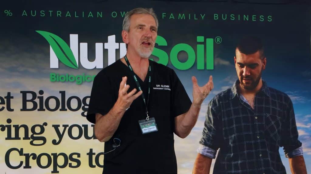 A man stands gesturing and speaking in front of a NutriSoil banner, which reads "Biological" and "Crops to" among other text. He has short grey hair, a beard, and is wearing a black shirt with an ID badge. Another person is partially visible on the banner behind him.