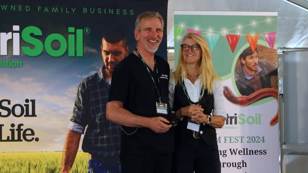 Two people stand smiling in front of banners promoting a business called "AgriSoil." The man is holding a microphone, and the woman stands beside him. The banners feature images of people and text about soil health and wellness.