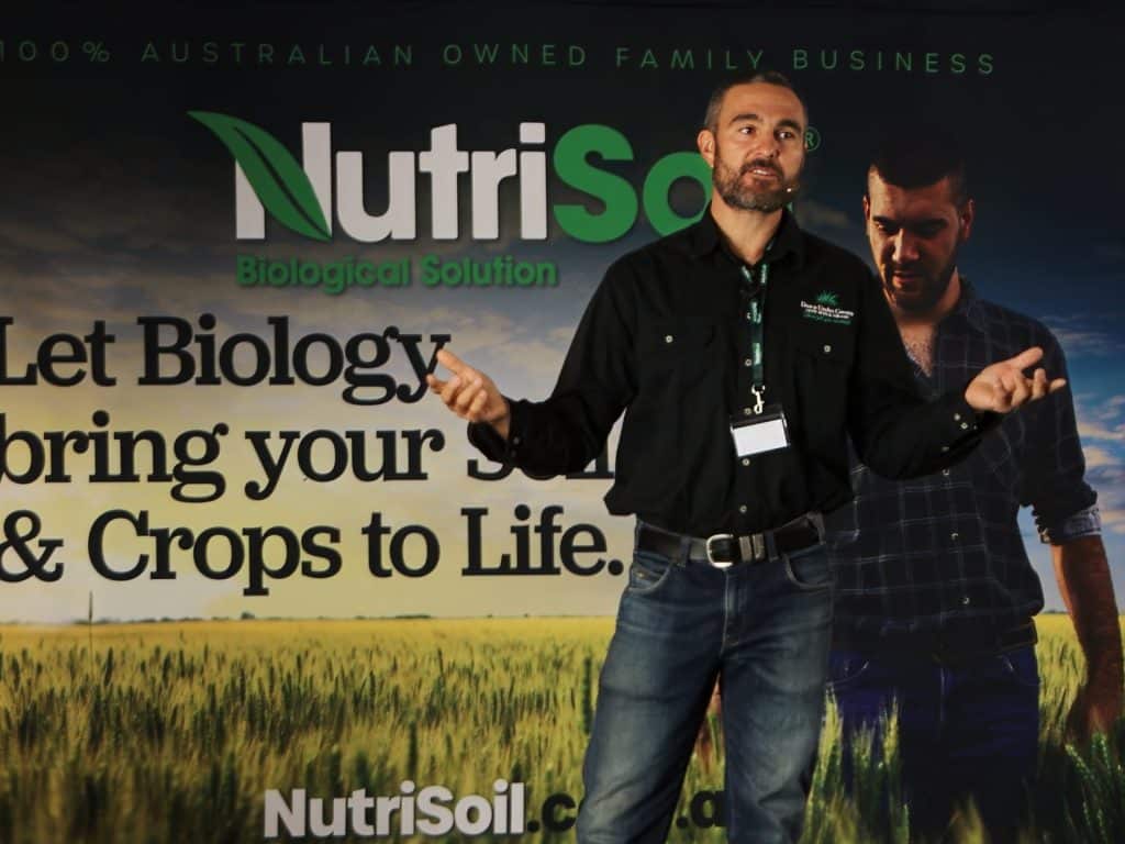 A man with a beard stands in front of a NutriSoil banner. He is wearing a black shirt and jeans, gesturing with both hands. The background displays the slogans "Let Biology bring your Soil & Crops to Life" and "100% Australian owned family business.