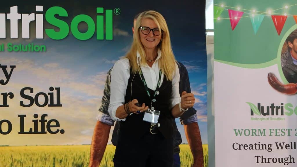 A woman wearing glasses and a professional outfit stands in front of a promotional backdrop for NutriSoil and NutriSoils Worm Fest. She is holding a presentation clicker and smiling, suggesting she is speaking at an event, possibly about soil health and biological solutions.