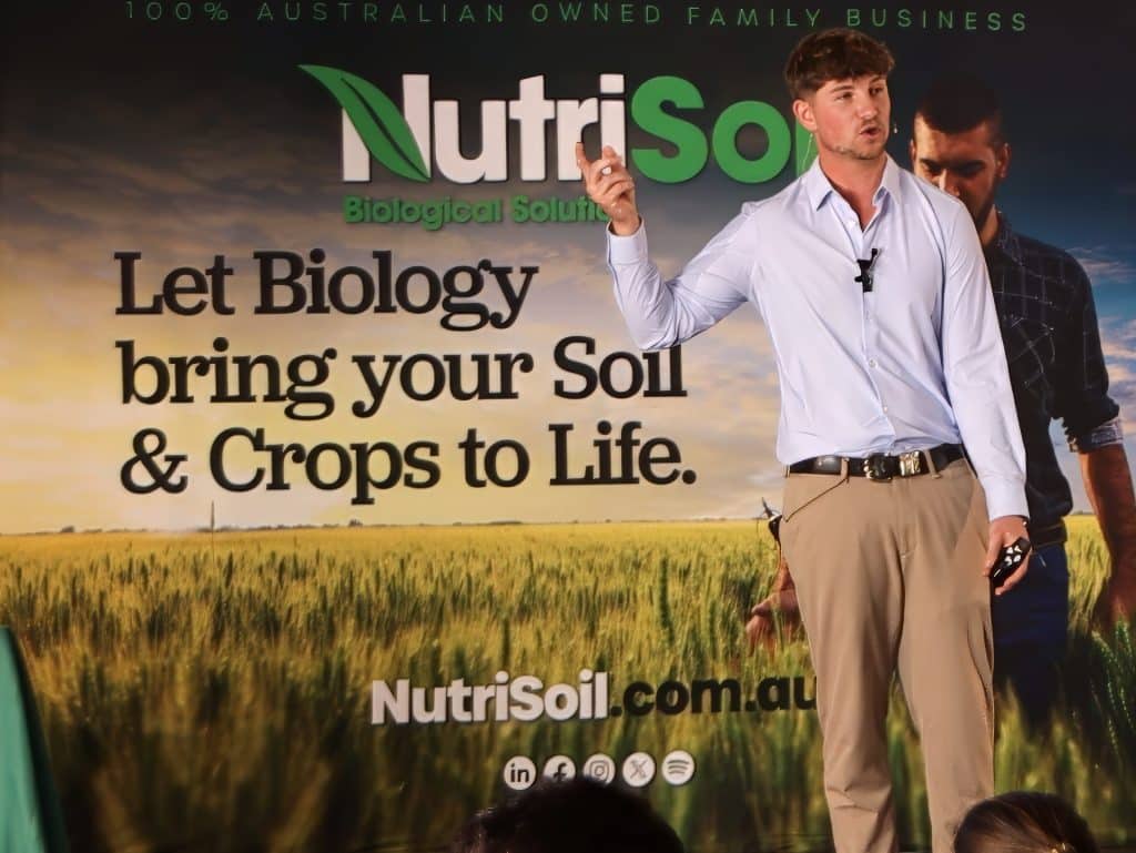 A man wearing a light blue shirt and khaki pants is speaking on a stage in front of a backdrop that reads "NutriSoil Biological Solutions. Let Biology bring your Soil & Crops to Life." The background features a green field and another person standing.