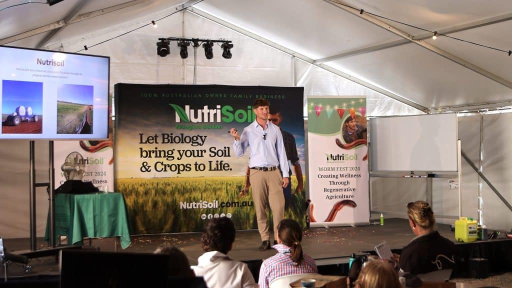 A speaker in a tent gives a presentation at the 2018 Nutrisoil Worm Fest. The backdrop reads "Let Biology bring your Soil & Crops to Life." Attendees sit at tables, and a screen and flipchart stand nearby. The event appears focused on sustainable agriculture.