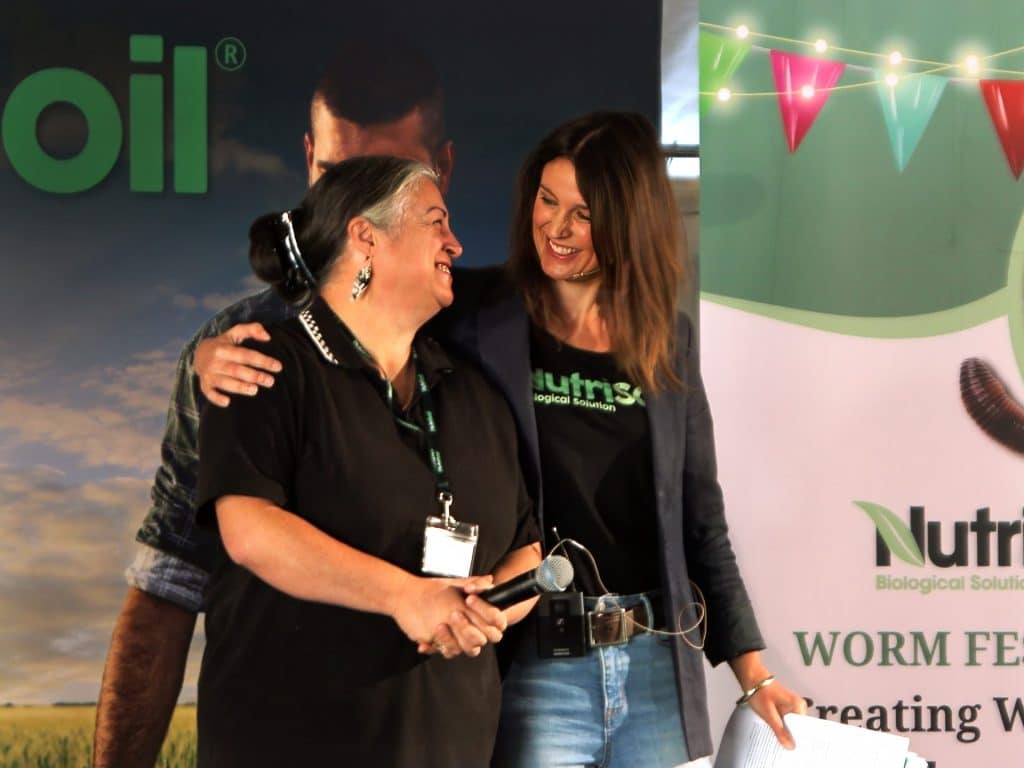 Two women stand closely, smiling warmly at each other. The woman on the left has gray hair and wears a black shirt, while the woman on the right has long brown hair and wears a blazer over a green shirt. They are speaking at an event; the banner behind them reads "Nutris" and "WORM".