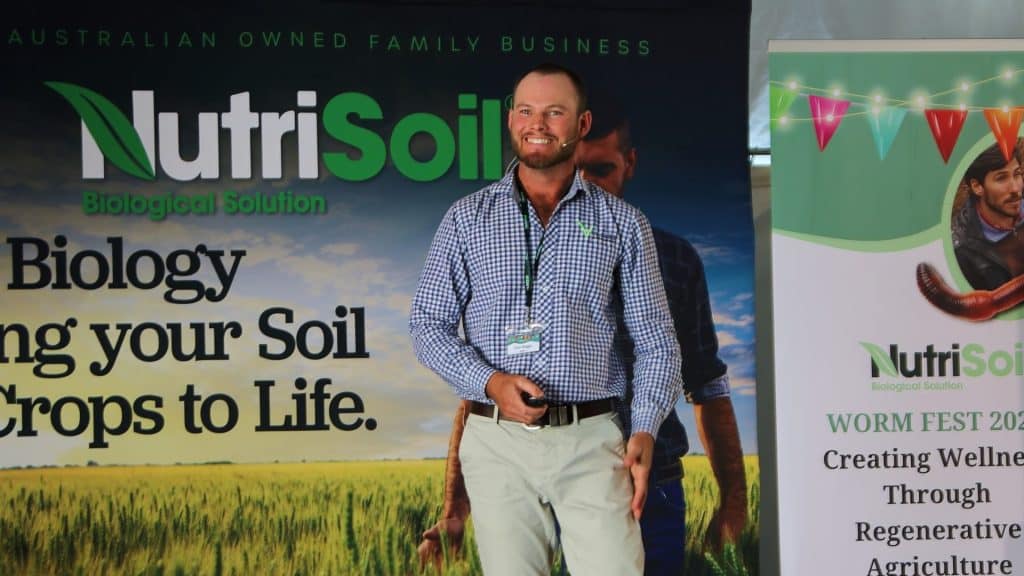 A man in a checkered shirt stands in front of a NutriSoil banner at WORM FEST 2022. The banner reads "NutriSoil Biological Solution: Bringing Your Soil and Crops to Life." He smiles and holds a microphone while giving a presentation on regenerative agriculture.