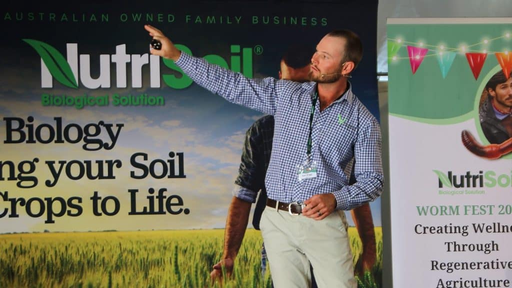 A speaker in a blue checkered shirt and gray pants, wearing a headset, is presenting on stage at an event. He is pointing to a slide on a screen. The backdrop displays the NutriSoil logo and a farming field. The event banner reads "WORM FEST 2023.
