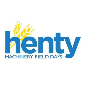 Logo with text "Henty Machinery Field Days" in blue. The word "henty" is prominently displayed, with two stylized yellow wheat stalks emerging from the 'h'. "MACHINERY FIELD DAYS" is written below in a smaller font. The background is white.