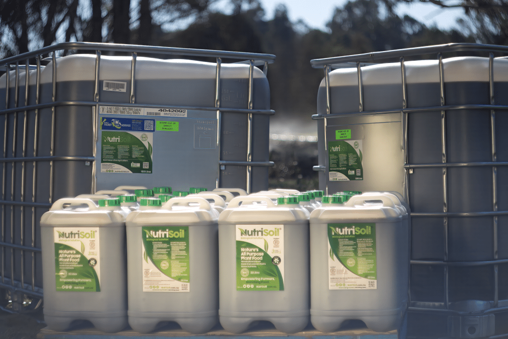 The image shows several containers and large plastic tanks with green labels, labeled "NutriSoil." The containers are situated outdoors on a sunny day, and trees can be seen in the background. There are two large tanks and multiple smaller canisters arranged on a pallet.