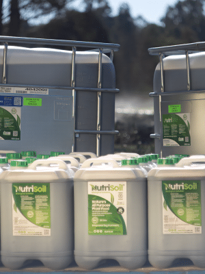 The image shows several containers and large plastic tanks with green labels, labeled "NutriSoil." The containers are situated outdoors on a sunny day, and trees can be seen in the background. There are two large tanks and multiple smaller canisters arranged on a pallet.